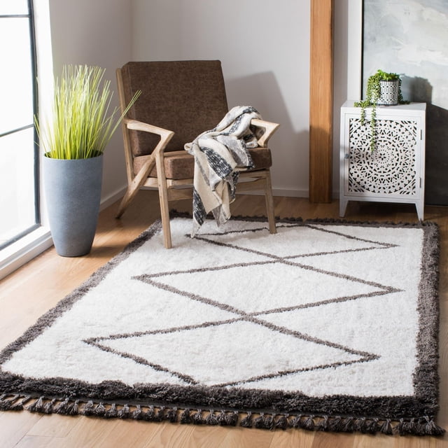 Safavieh Carmella Christopher Geometric Shag Area Rug featuring a white synthetic shag pile with dark brown geometric diamond patterns and a thick dark brown fringe border, placed on light hardwood flooring in a contemporary room with a wooden armchair draped with a black-and-white throw, a tall gray planter with green grass, and a white decorative cabinet with intricate cutout designs, ideal for adding texture and warmth to accent spaces.
