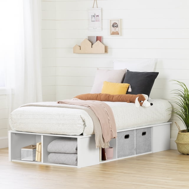 South Shore Flexible Twin Storage Platform Bed with Baskets in Pure White, featuring a low-profile frame with multiple open storage compartments and two grey fabric bins underneath. The bed is styled with a white textured mattress cover, pillows in black, beige, and yellow, and a long brown plush dog. A soft pink throw blanket is draped over the side. The room has white shiplap walls, minimalist wall art, and a woven basket with a green plant on a light wood floor. This bed is suitable for maximizing storage in small bedrooms.