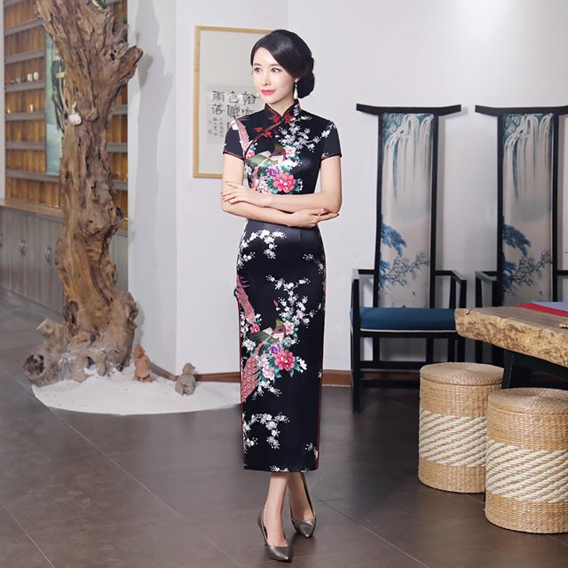 Robe Qipao Longue pour Femmes de style Rétro Classique Chinois Multicolore avec Motifs Floraux et Traditionnels. la Robe Ajustée Présente un Col Montant, des Manches Courtes et un Long Ourlet, Conçue pour un style Élégant et Intemporel. le Modèle Se Tient dans un Cadre Intérieur Traditionnel avec une Déco en Bois et Artistique, Portant la Robe Assortie à des Talons Argentés.