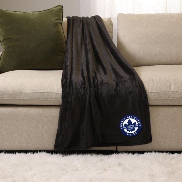 NHL, NBA, MLB Black Sports Throw Blanket, 50" x 60", draped over a beige sofa with a green pillow, made of soft plush 100% polyester, featuring a Toronto Maple Leafs logo patch in blue and white, suitable for indoor use and cozy warmth.