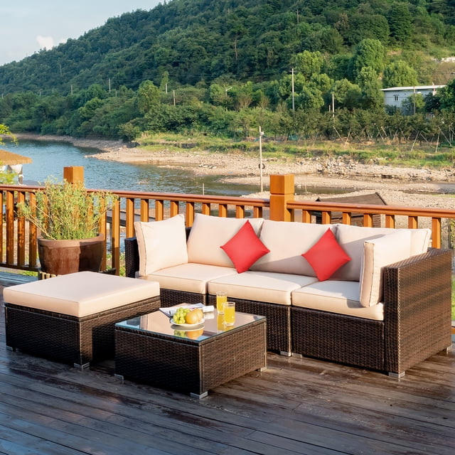 Costway 5PCS Outdoor Patio Rattan Furniture Set Sectional Conversation with beige cushions, featuring a modular design with 1 armless sofa, 2 corner sofas, 1 ottoman, and 1 coffee table with a tempered glass top. The set is crafted with a sturdy steel frame and weather-resistant PE rattan, suitable for outdoor use on patios, balconies, or gardens. The beige cushions are 4 inches thick, removable, and washable, with two red accent pillows included. The furniture is arranged on a wooden deck overlooking a scenic riverside view.