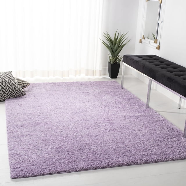 SAFAVIEH August Carlene Solid Plush Shag Area Rug in soft lavender purple with a thick polypropylene pile, displayed on a white floor in a bright room with sheer curtains, a black tufted bench with chrome legs, a green potted plant, and two black and white patterned pillows, ideal as a plush accent rug for modern interiors.