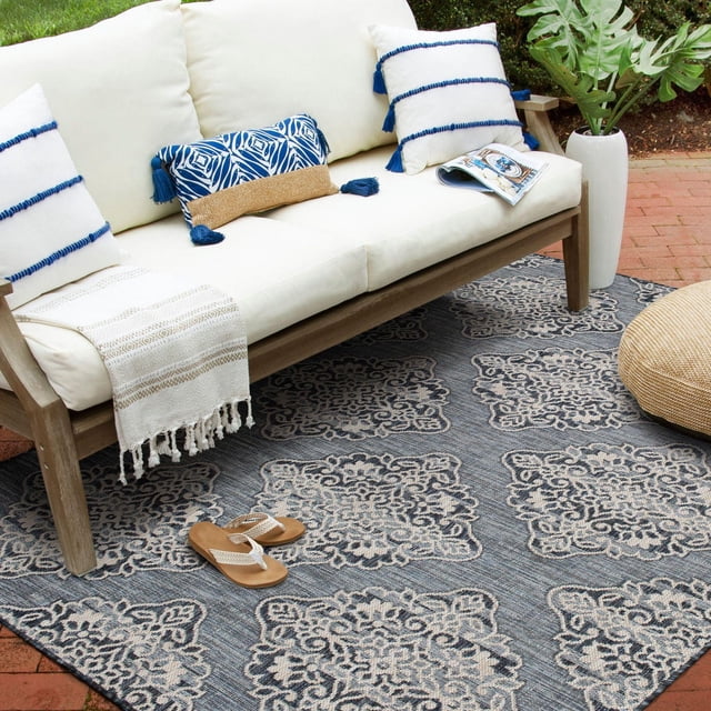 Hayweld Lagoon Blue and White Woven Area Rug featuring a diamond trellis and ornate white pattern on a slate blue background, set on a brick patio under a cream cushioned wooden sofa with blue and white tassel pillows, alongside a beige woven pouf and a white vase with green leaves, suitable for indoor or outdoor spaces.