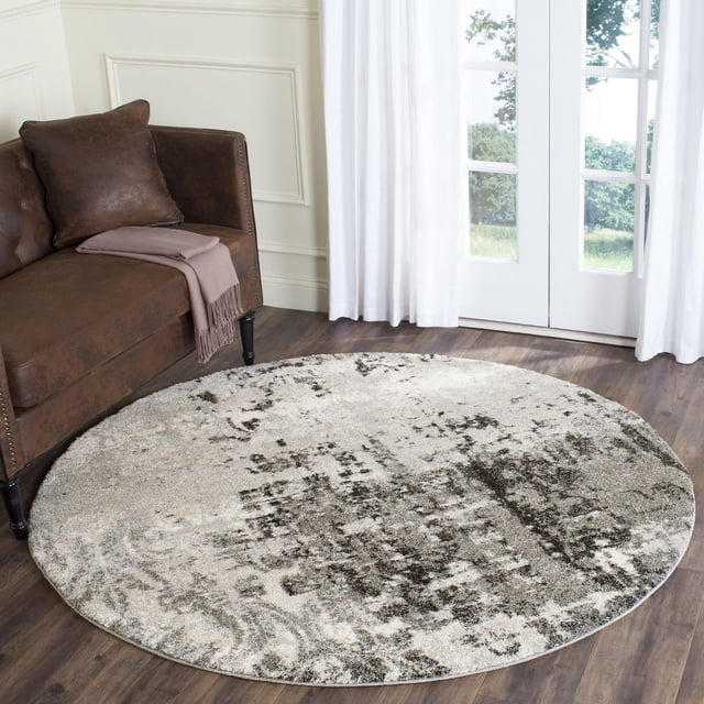 Safavieh Retro Corynn Abstract Area Rug, a round low-pile polypropylene rug with a 1960s mid-century modern abstract pattern in beige and dark gray, placed on hardwood flooring beside a brown leather sofa with a mauve throw, near white curtains and glass doors, ideal for accenting living rooms.
