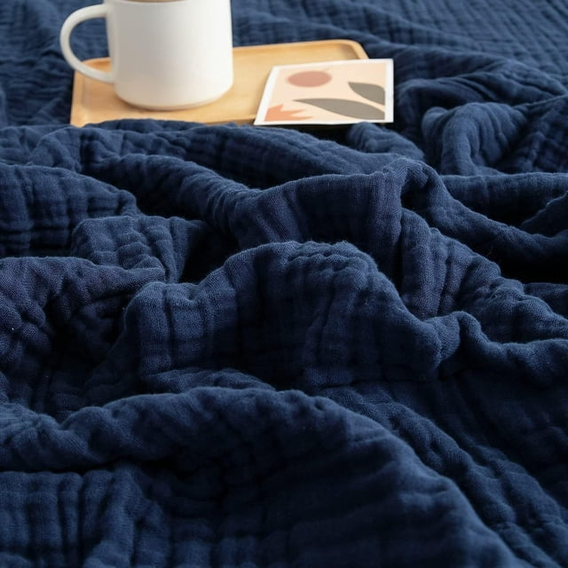 EGNMCR Couverture Jetée, une Couverture en Mousseline Douce de Couleur Bleu Marine Foncé, Parfaite pour Canapés, Lits Ou comme Couverture d'Été Légère. Fabriquée en Tissu Mousseline Respirant, Elle Régule Naturellement la Température et Offre un Confort Doux comme un Nuage pour une Utilisation Toute l'Année. la Couverture Est Exposée avec des Plis Texturés, à Côté d'Un Plateau en Bois Tenant une Tasse Blanche et une Carte Décorative, Soulignant Son design Polyvalent et Confortable.