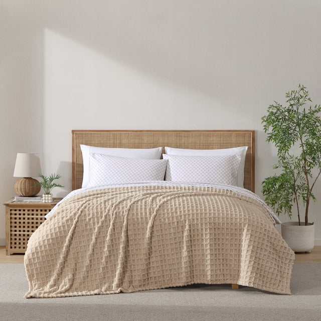C&C California Jacquard Sherpa Blanket in oat with a textured windowpane grid weave, laid over a bed with a woven rattan headboard, styled with white patterned pillows, a wooden nightstand holding a lamp and small plant, and a potted green plant on a grey carpet in a neutral bedroom, ideal for all-season comfort and versatile use.