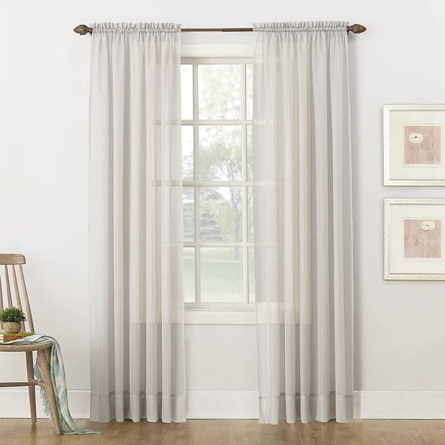 SUN TEXTILE VOILE ROD POCKET CURTAIN in sheer voile fabric with built-in rod pockets, hanging on a decorative rod with finials, gently filtering natural light through a window in a bright room with wood flooring, a wooden chair with a plant and scarf, and framed floral prints, ideal for layering sheer curtains to enhance light control and privacy.