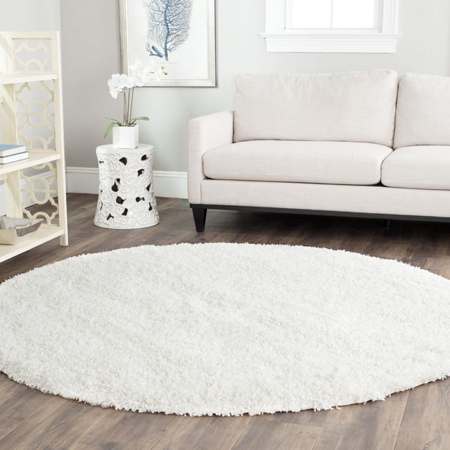 Safavieh California Tapis Shag en Peluche Uni, Rond avec un Épais Velours de Polypropylène Blanc Pesant 4,5 kg par Mètre Carré, Exposé sur un Plancher de Bois Franc Foncé dans un Salon avec un sofa beige, une table d'Appoint en Céramique Blanche et des Étagères en Bois Clair, Convient pour Ajouter du Confort en Peluche et un style Décontracté de la Côte Ouest.