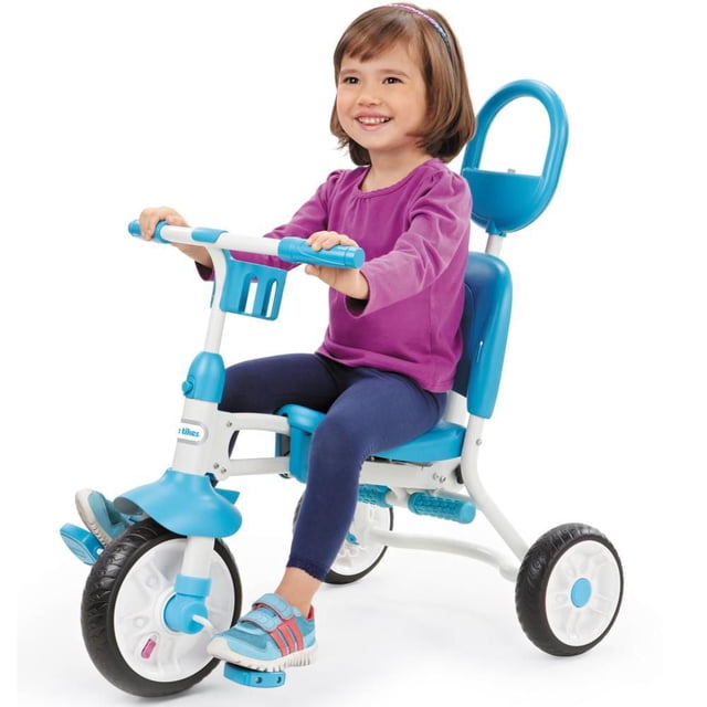 Little Tikes Pack ‘n Go Trike with a child seated, featuring a white and blue folding tricycle frame, large front wheel with textured black tire, two smaller rear wheels, blue seat with backrest, foldable footrests, and blue handlebars with grips and a small front basket for portability.