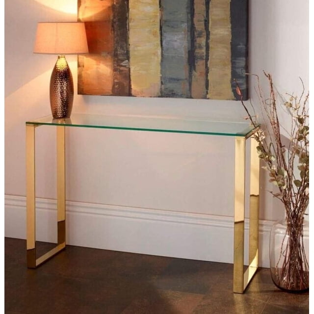 Plata Import Gold Gen Console Medium with a clear tempered glass tabletop and gold stainless steel legs, placed in a living room setting with a textured lamp on the left and a tall vase with dried branches on the right beneath modern abstract wall art, suitable as an accent table or display surface.