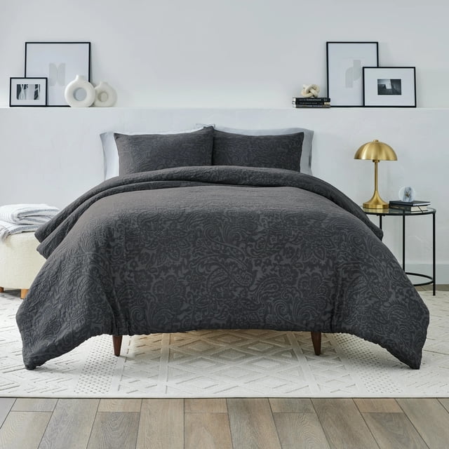Better Homes & Gardens Comforter Set in grey with a subtle floral damask pattern, crafted from a breathable cotton blend. Includes a comforter and two pillow shams with envelope closures, displayed on a wooden bed frame in a minimalist bedroom with white walls, framed abstract art, a gold table lamp, and a textured white area rug on hardwood flooring. Ideal for all-season comfort and adding soft texture to bedroom décor.