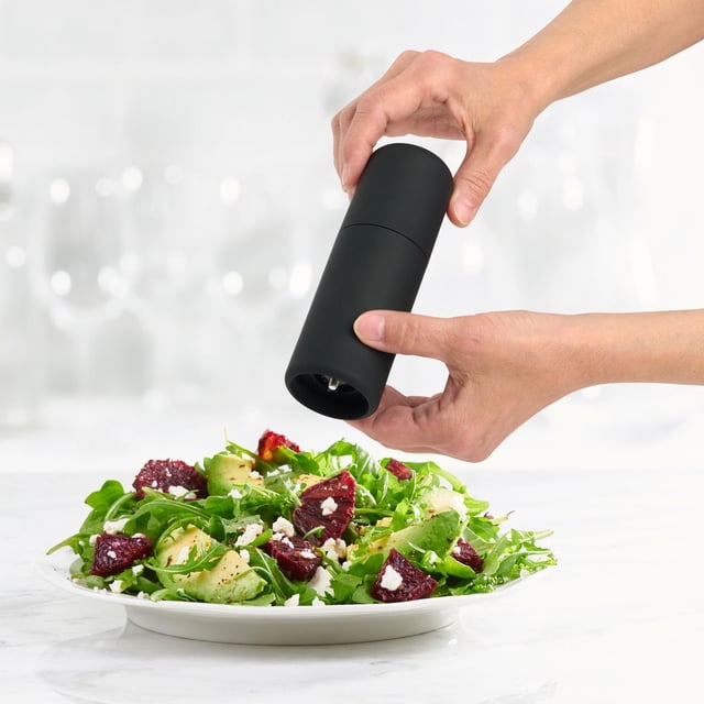 Trudeau 5.5" Lexi Salt and Pepper Mills in matte black, unfilled, shown held above a fresh salad with greens, avocado, and beetroot, featuring a durable ceramic grinding mechanism and compact design suitable for everyday meals and entertaining.