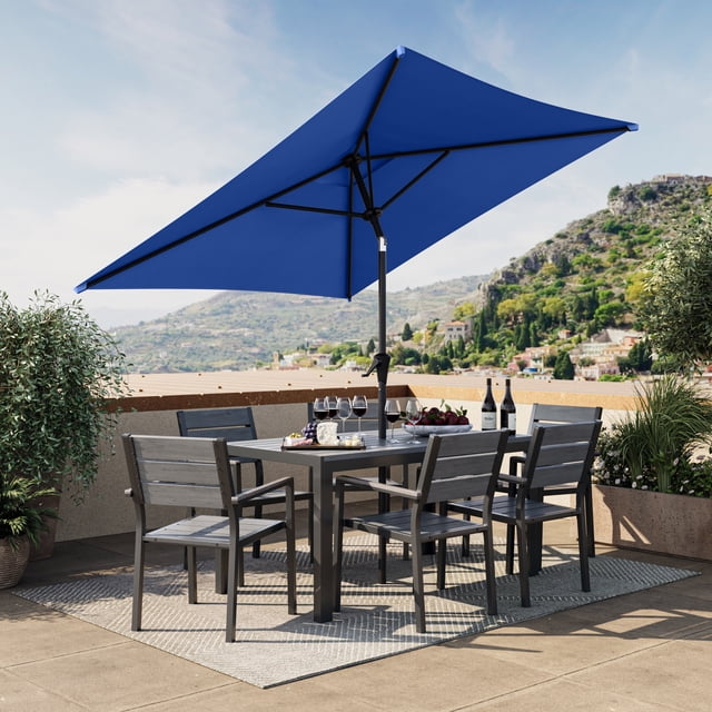 CorLiving 6.5ft x 6.5ft Square Tilting Outdoor Market Umbrella with steel frame and cobalt blue canopy shading a patio dining set with six grey metal chairs and a square table on a tiled terrace with mountain views, featuring a vented canopy for airflow and an easy tilt mechanism for adjustable shade.