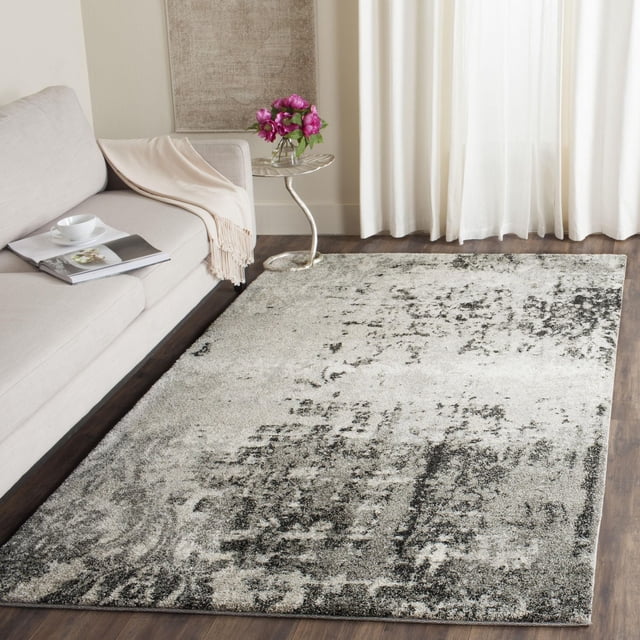Safavieh Retro Corynn Abstract Area Rug featuring a low pile polypropylene construction with a mid-century modern abstract pattern in grey and black on a light background, placed on dark hardwood flooring beside a beige sofa with a cream throw and a silver side table with pink flowers, suitable for enhancing contemporary living rooms.