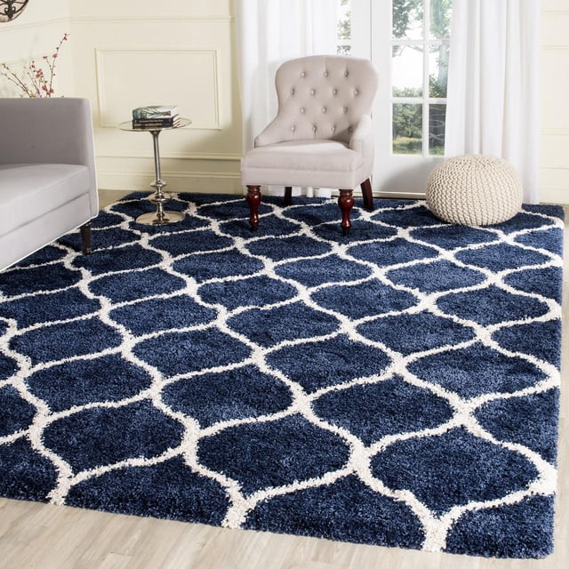 Safavieh Hudson Tapis Shag Géométrique Arline avec une Épaisse Peluche Shag Bleu Marine et des Motifs de Carreaux en Forme d'Oignon Blanc, Exposé dans un Salon avec un Fauteuil Capitonné beige, une table d'Appoint en Métal avec des Livres, et un pouf Tricoté Crème Près d'Une Porte en Verre avec des Rideaux Blancs, Convient pour Ajouter de la texture aux Espaces à Fort Passage.