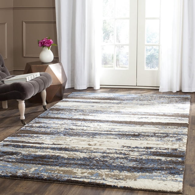 SAFAVIEH Retro Mayson Abstract Area Rug in a bright room with hardwood floors, featuring a low pile polypropylene surface with horizontal abstract stripes in blue, beige, and cream tones, styled with mid-century modern furniture including a tufted gray chair and geometric side table with a white vase holding pink flowers, ideal for accenting living areas.