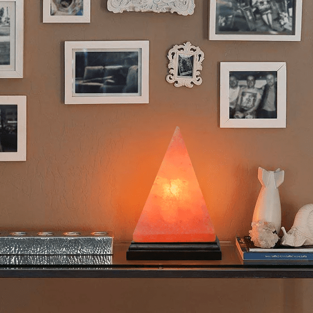 Lampe en Forme de Pyramide Rose Artisanale Himalayenne par Apex Global, avec une Lumière Ambre Lumineuse. la Lampe en Forme de Pyramide Est Fabriquée à Partir de Sel Rose de l'Himalaya durable et Est Exposée sur une base Noire. Positionnée sur une Étagère dans une Pièce Douillette avec des photos Encadrées et des Articles Décoratifs, Elle Sert d'Idée-Cadeau unique et de Pièce d'Éclairage Fantaisie Élégante.