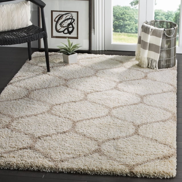 Safavieh Hudson Tapis Shag Géométrique Arline avec une Épaisse pile Shag Ivoire et un Motif de Carreaux Géométriques Brun, Exposé sur un Plancher de Bois Franc Foncé. le Tapis Est Conçu avec une Chaise Tressée Noire, un Coussin Crème, une Petite Plante en Pot et un Panier en Fil Tenant un Jeté Rayé Près de Portes en Verre Surplombant la Verdure. Idéal pour Ajouter de la texture et du Confort aux Espaces de Vie Modernes.
