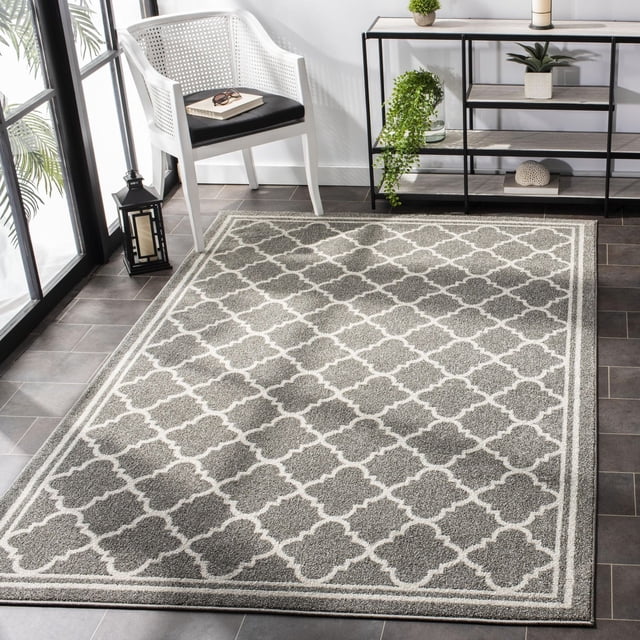 SAFAVIEH Amherst Trina Geometric Area Rug with grey and white quatrefoil pattern, power-loomed polypropylene for durable indoor-outdoor use, displayed on dark tile floor near white woven armchair and black metal shelving with plants, suitable for high-traffic areas.