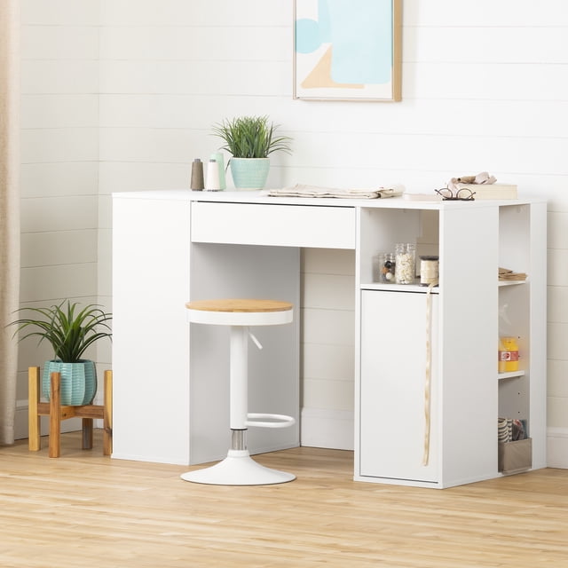 South Shore Crea Counter-Height Craft Table in pure white with a central drawer, open storage compartments, and a cabinet door on the right side, ideal for office furniture and craft workspace. Paired with a white adjustable stool with a round wooden seat. The table surface holds a small potted plant, spools of thread, folded fabric, and a book with glasses. The setting features light wood flooring, white shiplap walls, and a framed abstract artwork above.