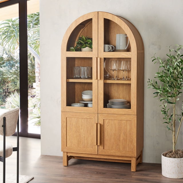 Better Homes & Gardens Juliet Kitchen Rounded Arch Cabinet in light honey finish with rounded arch top, two tempered glass doors displaying glassware, plates, and a plant on adjustable shelves, and two solid wood doors below for concealed storage, standing on short legs against a light wall near a window and potted plant, suitable for kitchen or dining room storage.