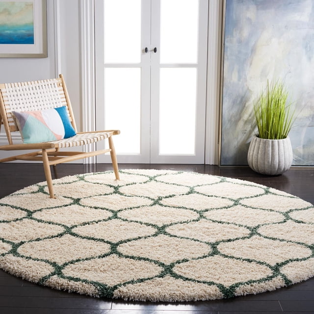 Safavieh Hudson Arline Geometric Shag Area Rug, round with a thick ivory shag pile and green geometric tile pattern, placed on dark hardwood flooring beside a woven wooden chair with a multicolored cushion and a tall potted plant, suitable for adding texture to modern living spaces.