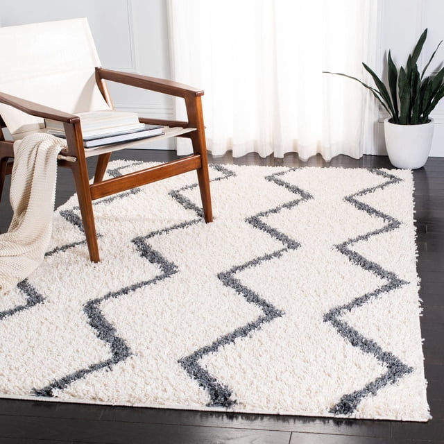 Safavieh Venus Carleen Chevron Shag Area Rug with a 1.77-inch medium pile in white and dark gray chevron pattern, placed on dark hardwood flooring beside a mid-century modern wooden armchair with cream fabric and beige knitted throw, near sheer white curtains and a potted snake plant, suitable for adding plush texture and warmth to contemporary living spaces.