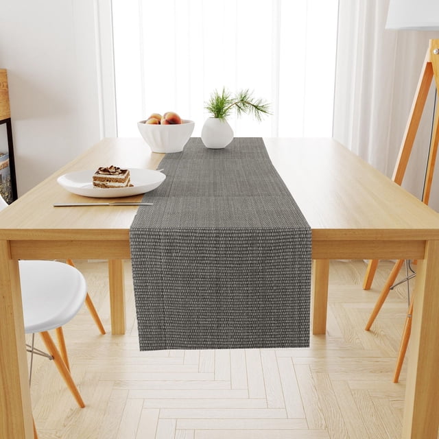 Fabstyles Casual Classic Cotton Table Runner in dark gray woven cotton fabric measuring 13x54 inches, centered on a light wood dining table with a white plate holding a slice of layered cake and fork, a white bowl with peaches, and a small white vase with pine sprigs, in a bright room with light wood herringbone flooring and white curtains, also suitable as a coffee or patio table runner.