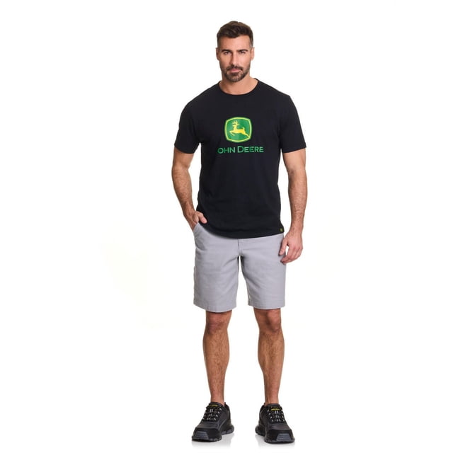 John Deere Short de Charpentier en Canevas Extensible Gris Pâle pour Homme, Porté par un Mannequin Debout avec les Mains dans les Poches, Assorti à un t-shirt logo John Deere Noir et à des Chaussures Athlétiques Noires, Offrant une Coupe Longueur Genou et une construction durable Adaptée aux Vêtements de Travail.