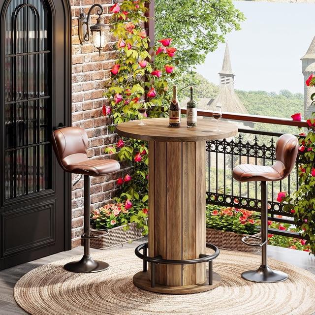 39.4-inch high wood bar table with a warm-brown wood-grain round tabletop and a distinctive fence-style wooden base, featuring a metal ring footrest. The table is displayed on a balcony with two brown leather bar stools, surrounded by vibrant flowers and a scenic outdoor view. Its compact circular design, with a 31.1-inch diameter, is ideal for small spaces like apartments or cafes, blending farmhouse and industrial styles.