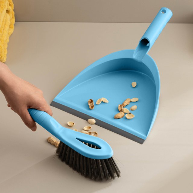 RKSTN Broom with Dustpan Combo Set featuring a compact blue dustpan and matching handheld brush with black bristles. The set is designed for cleaning small spaces like desktops, sofas, and car interiors. The image shows the brush sweeping peanut shells into the dustpan on a beige surface. The lightweight plastic construction and ergonomic handles make it easy to use and store.