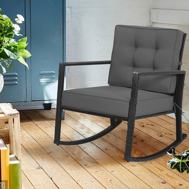 Costway Patio Rattan Rocker Chair Outdoor Glider Rocking Chair Cushion Lawn Grey. A black rattan rocking chair with grey tufted cushions, designed for comfort with a thick seat cushion and angled backrest. Suitable for indoor or outdoor use, including nurseries, patios, or living rooms. The chair is shown on a wooden floor in a rustic indoor space with plants and lockers in the background.