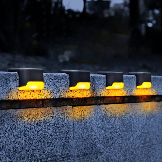 4pcs LED Solar Lights Deck Lights by LIVINGbasics, shown installed on a stone wall at night. These waterproof, warm light outdoor solar wall lights automatically turn on at night and off at dawn. Ideal for decks, patios, stairs, yards, paths, and driveways. The lights feature a durable ABS construction, IP45 waterproof rating, and a polysilicon solar panel that charges in 6-8 hours for 8-10 hours of illumination. Includes screws and double-sided adhesive for easy mounting.