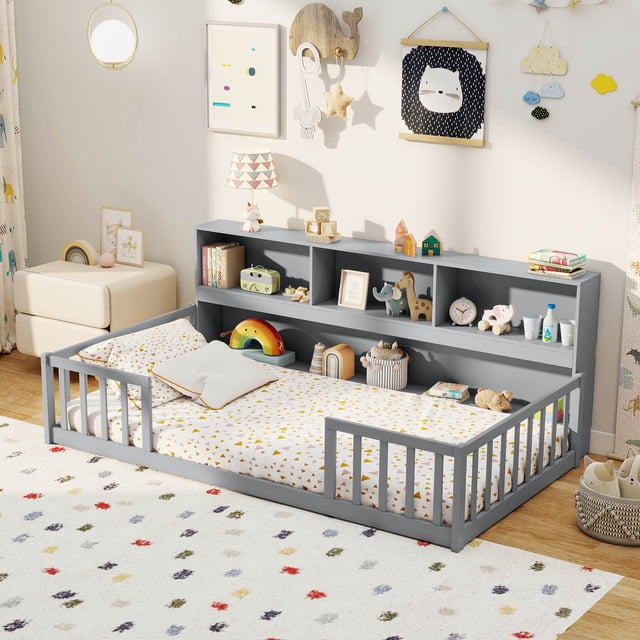 Costway Twin Size Montessori Floor Bed with Bookcase Storage Shelves & Safety Fence Rails in gray. The bed features a low-to-ground design with full-length safety fence rails and a wide entrance for easy access. Adjacent to the bed is an integrated bookshelf with multiple compartments, ideal for organizing books, toys, and learning materials. Crafted from solid pine wood and engineered wood, this bed promotes independence and serves as both a sleeping space and a creative play area for children. The setup is displayed in a bright, playful children's room with colorful decor and toys.