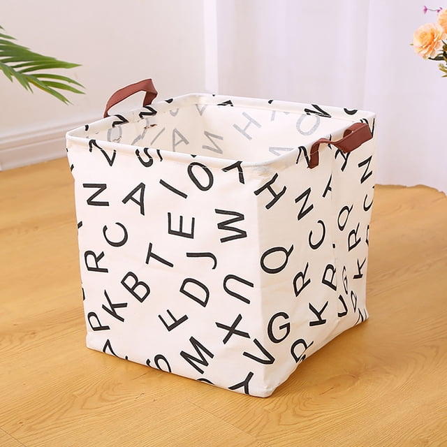 Laundry Basket Dorm Baby Laundry Basket Large Laundry Hamper with Handle for Dirty Clothes, featuring a foldable fabric design with black alphabet print on a white background. Includes sturdy brown handles for portability and a spacious interior for organizing laundry, toys, or household items. Perfect for laundry room storage, displayed on a wooden floor in a modern, minimalist room.
