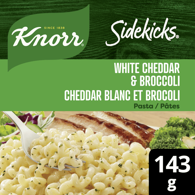 Knorr Sidekicks White Cheddar & Broccoli Pasta Side Dish showing creamy white cheddar sauce with broccoli florets mixed into spiral pasta, a fork lifting pasta, grilled meat and fresh broccoli in the background, packaged weight 143 g, suitable for quick, easy family meals or as a standalone side dish.