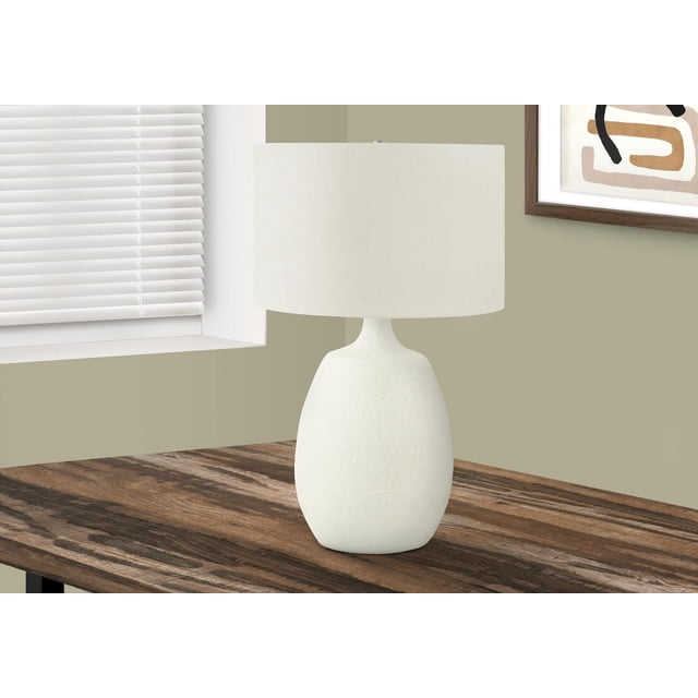 Monarch Specialties Lighting 26" h table lamp with a textured cream resin base and ivory linen shade on a dark wood side table near a window with white blinds and abstract framed art, suitable for living room or office ambient lighting.