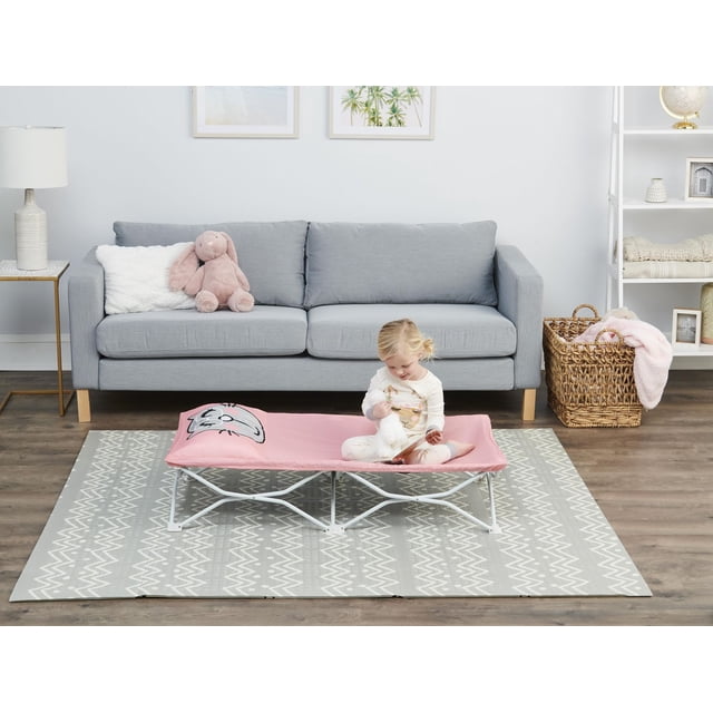 Regalo My Cot Pal Pink Kitty portable toddler bed with pink kitty face fabric, set on a grey geometric patterned rug in front of a light grey sofa with a pink plush bunny and white pillow, a toddler seated on the cot holding a book and stuffed animal, with a woven basket and white shelving unit in a modern living room.