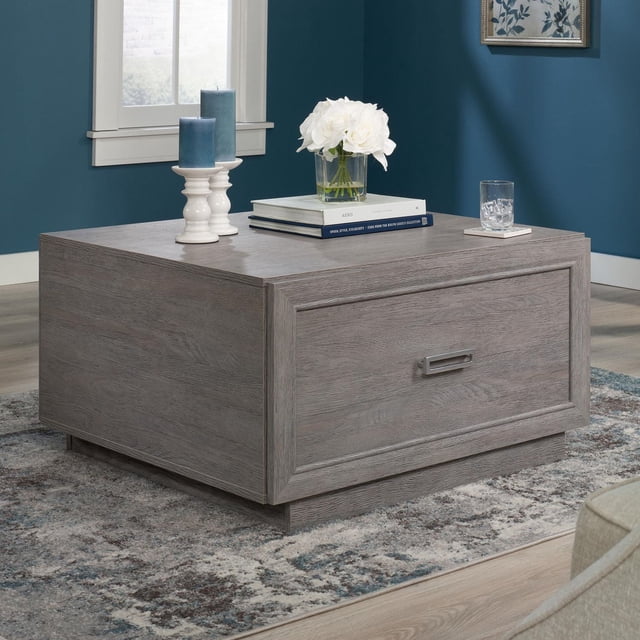 Sauder Hayes Garden Table Basse en Chêne Cendré au design Carré, avec un Grand Tiroir à Glissières à extension Complète et une Poignée Métallique Géométrique, plus une Tablette Ouverte pour le Rangement, Placée sur un Tapis à Motifs dans un Salon aux Murs Bleus, Décorée de Bougies Blanches, de Livres et d'Un vase en Verre de Fleurs Blanches. Elle Convient Aussi pour Organiser des Couvertures Ou des Accessoires Multimédias.