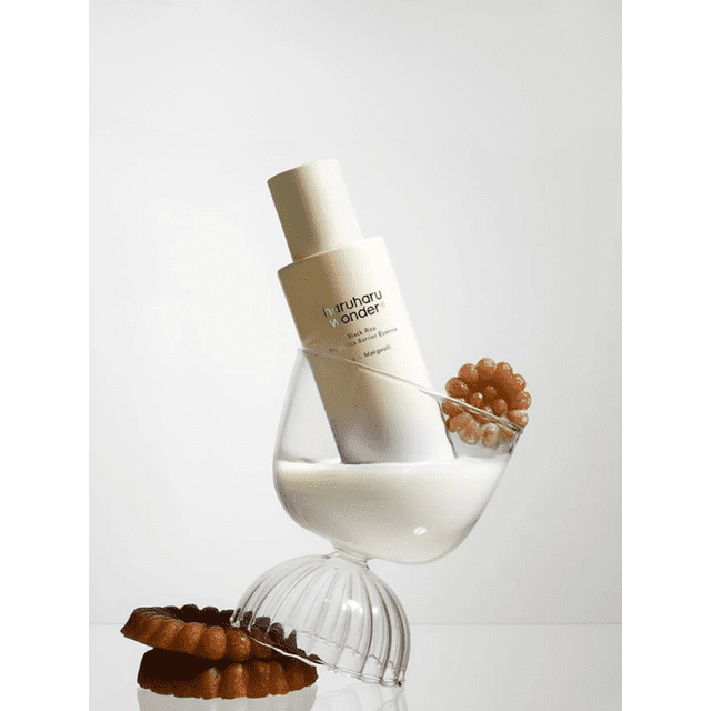 haruharu wonder Black Rice Probiotics Barrier Essence 120ml bottle angled inside a clear glass bowl half-filled with white liquid, accompanied by two brown ring-shaped textured objects, set against a plain white background, emphasizing its lightweight, hydrating formula that strengthens the skin barrier.