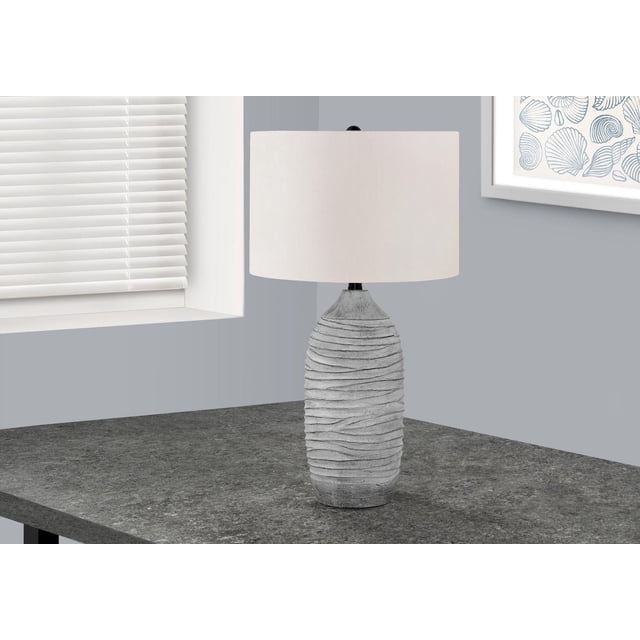 Monarch Specialties Lighting 27"h table lamp with a textured grey resin vase-shaped base featuring flowing lines, an ivory linen drum shade, and a black metal finial, placed on a dark grey tabletop near a window with white blinds and framed shell artwork on a grey wall, ideal for modern living rooms or bedrooms.