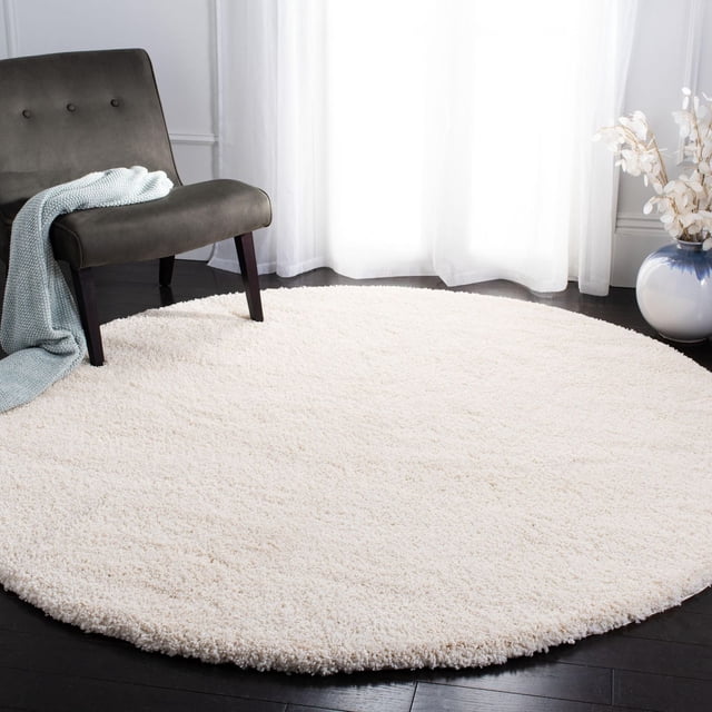 Safavieh California Solid Plush Shag Area Rug, round with a dense 4.5kg/m² polypropylene pile in cream, placed on dark hardwood flooring beside a dark grey upholstered chair with a light blue knitted throw, near sheer white curtains and a blue and white ceramic vase with dried flowers, suitable for adding softness and warmth to living spaces.