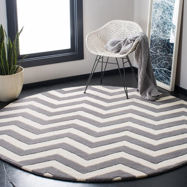 Safavieh Chatham Jake Tapis à Rayures Zigzag de Forme Ronde et Rayures Zigzag Grises et Blanches, Velours de Laine Tufté à la Main pour une Douceur de Hauteur Moyenne, Posé sur un Plancher Sombre Près d'Une Chaise Tissée Blanche avec des Pattes en Métal Noir et un Jeté Gris, à Côté d'Une Plante Serpent et d'Une Oeuvre Encadrée Abstraite.