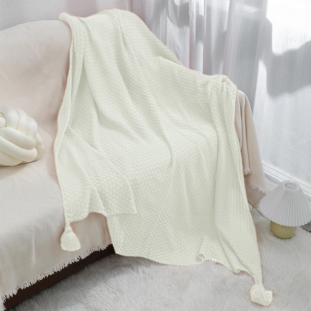 Acrylic knitted sofa blanket with wind fringe in white, measuring 100x70x0.5cm, draped over a beige sofa in a cozy living room setting. The blanket features a textured weave and tassel accents, ideal as a throw blanket or bedding accessory for office, nap, and home use.