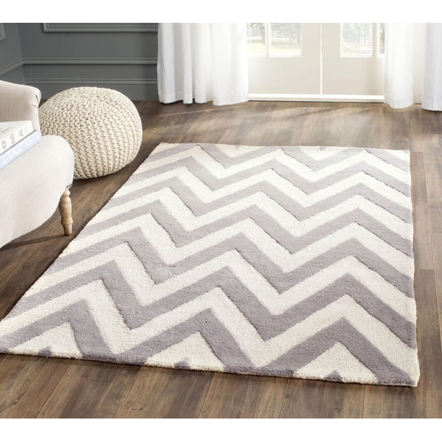 Safavieh Cambridge Kaitlyn Zig Zag Stripes Area Rug with hand-tufted wool in light grey and off-white zig zag stripes, shown on hardwood flooring in a bright room with a beige armchair and knitted pouf, ideal for adding texture and warmth to home or office spaces.