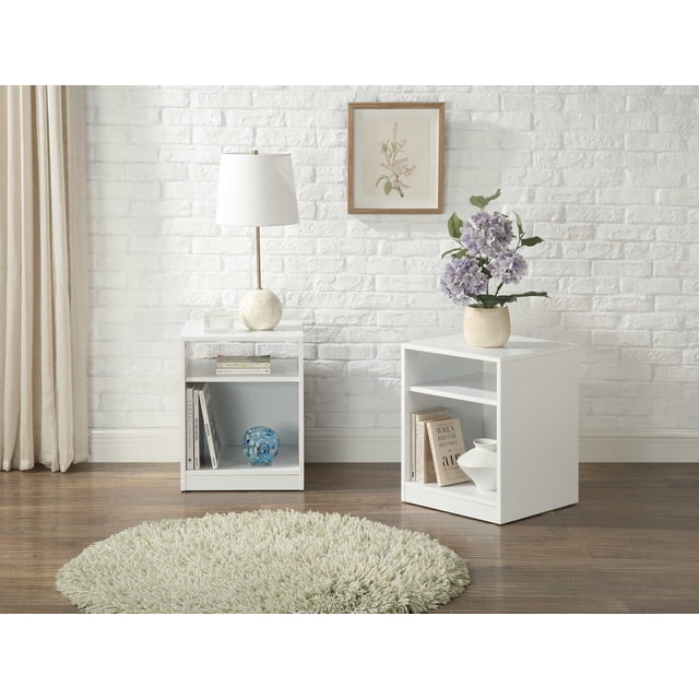 Mainstays Open Shelf Nightstand, Set of Two, White, shown in a room with white brick wall and wood floor. Each bedside table features a square shape with an open top shelf and a large lower cubby holding books and decorative items. One nightstand has a white lamp and glass container on top, the other a beige vase with purple flowers. A round cream shag rug is in front, emphasizing its open storage design.