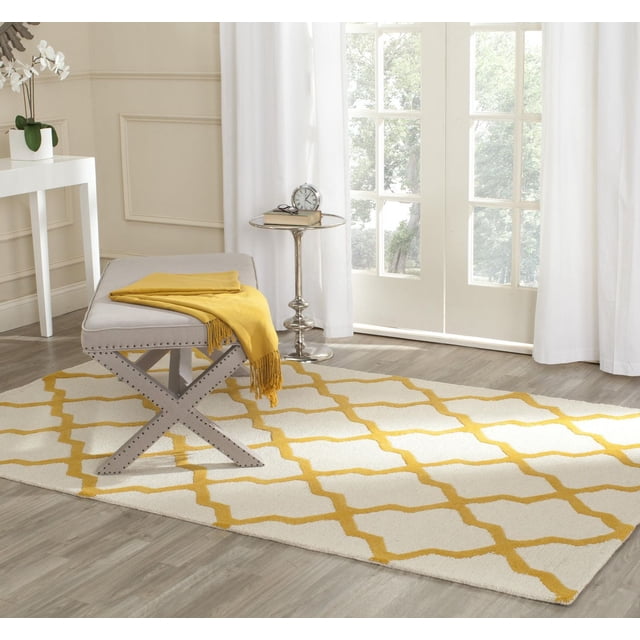 Safavieh Cambridge Liam Tapis Géométrique pour Aire avec une base Crème avec un Motif de Treillis Géométrique Jaune, texture de Laine Tuftée à la Main, Placé sur un Plancher de Bois Franc Clair sous un Banc Rembourré Gris avec Garniture de Clous Décoratifs et un Jeté Jaune, à Côté d'Une table d'Appoint Argent Poli avec une Horloge et un Livre, Rideaux Blancs Encadrant des Portes en Verre dans une Pièce Lumineuse.