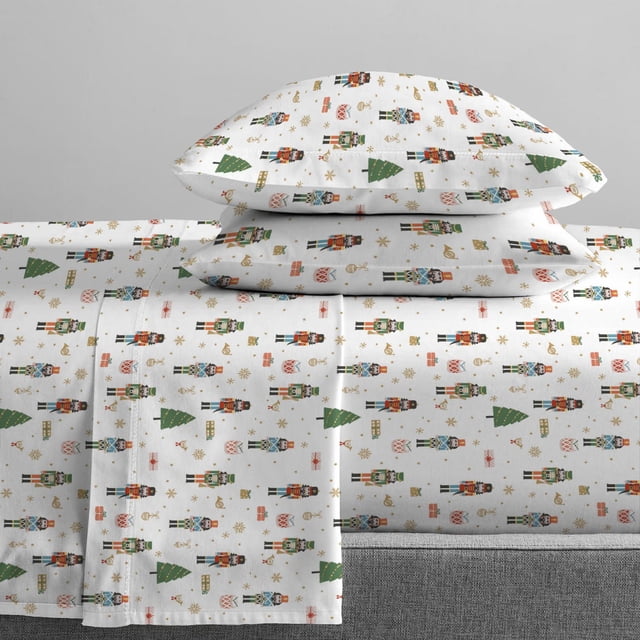 Johnson Home 144 Thread Count Cotton Printed Sheet Sets in white percale cotton featuring festive nutcracker soldiers, green Christmas trees, red drums, and gold snowflakes, with deep 15-inch pocket fitted sheets and matching pillowcases suitable for holiday bedroom styling.
