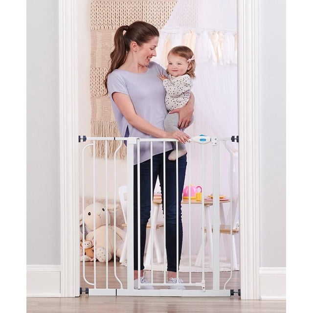 Regalo Extra Tall Walk Through Baby Gate in white, pressure-mounted in a doorway, standing 36 inches tall and fitting openings 29 to 38.5 inches wide, with an easy slide latch that locks automatically and a walk-through door, shown with a woman holding a toddler behind it in a softly decorated room with toys and a small table.