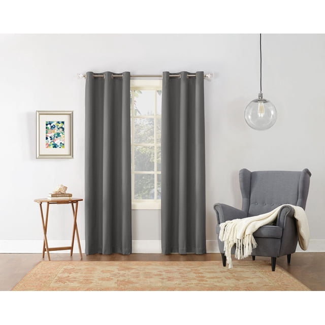 MAINSTAYS Textured Thermal-Lined Grommet Window Curtains Panel in dark grey with woven texture and thermal blackout lining, hanging on a silver metal rod over a window in a modern living room with light grey walls, a wooden side table with books, a grey wingback chair with a cream throw, and a clear glass pendant light. The thermal lining reduces energy loss and noise, enhancing room comfort.